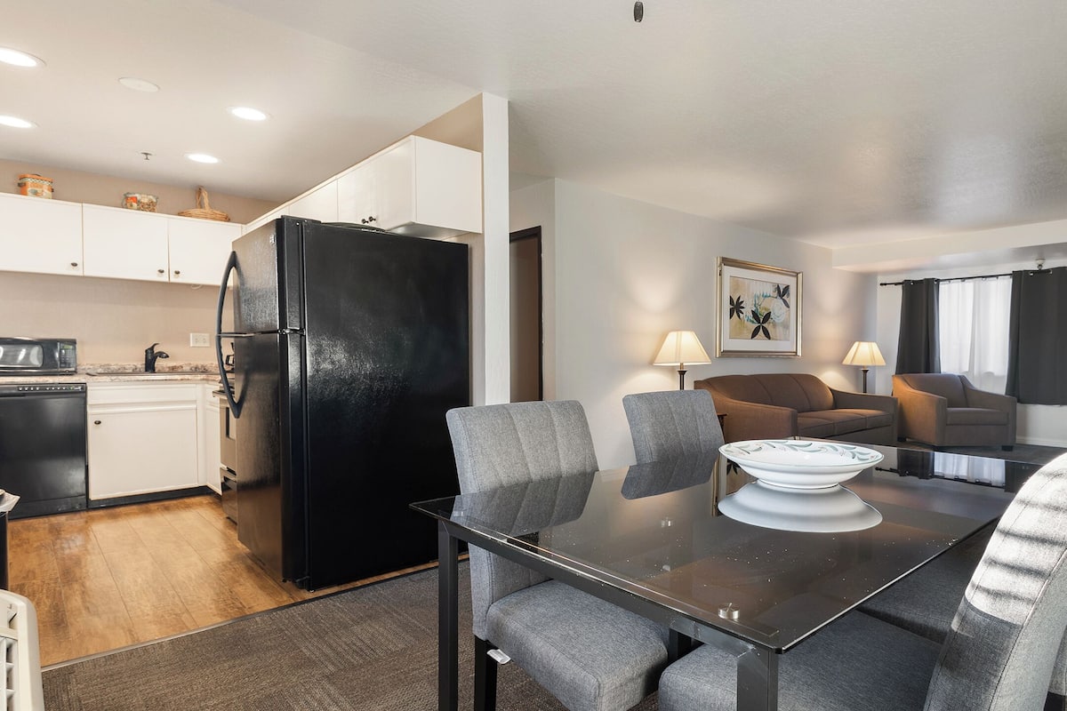 A compact kitchen with a black refrigerator, white cabinets, and a microwave. There is a dining area with a table, four grey chairs, and a white bowl. A brown sofa and two lamps are in the living area.