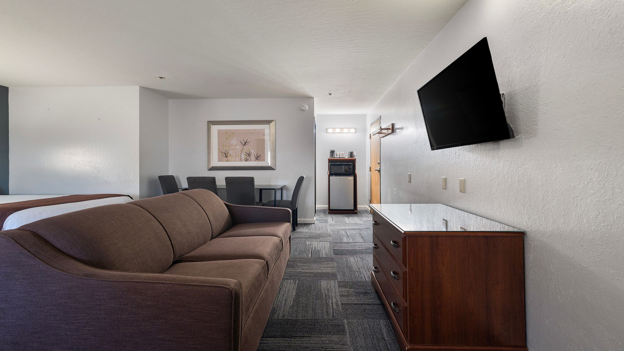 Ein Hotelzimmer mit einer Couch, einem an der Wand montierten Fernseher, einer kleinen Küchenzeile und einem gerahmten Bild.