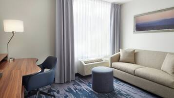 Habitación de hotel con un escritorio de madera, una silla de oficina azul, un sofá beige, un reposapiés gris y un ventanal con cortinas de gasa.