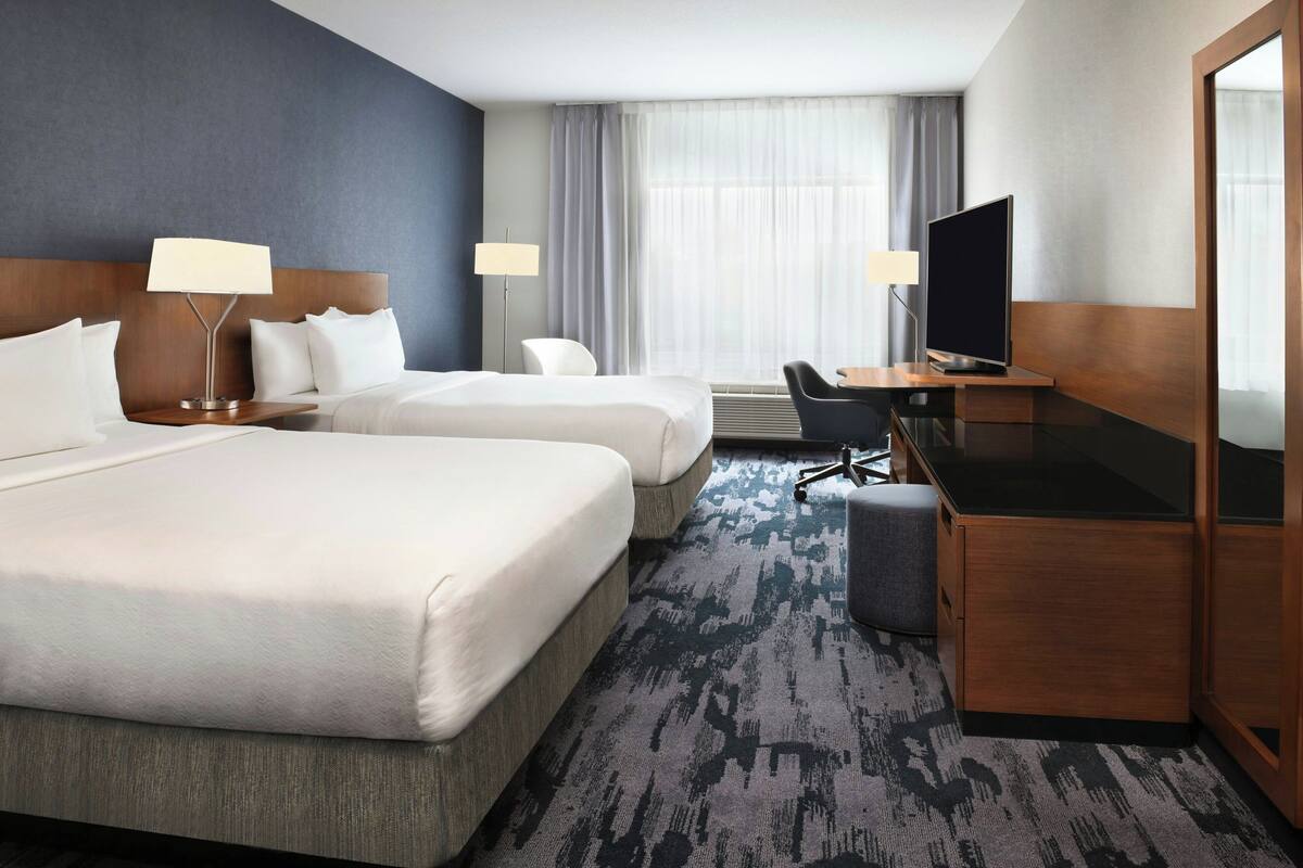 A hotel room with two beds, a desk, a TV, and a large window with curtains.
