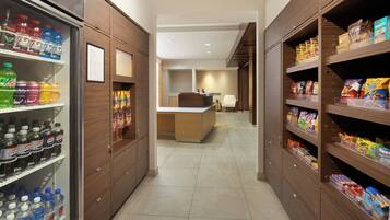 Un pasillo de hotel con un refrigerador lleno de bebidas, una kitchenette y estantes con aperitivos y artículos de aseo.