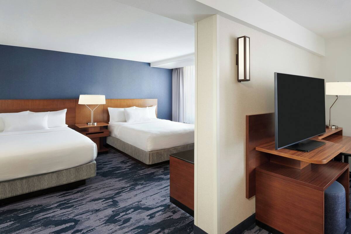 A hotel room with two beds, a desk with a flat-screen TV, a lamp, and a large window.