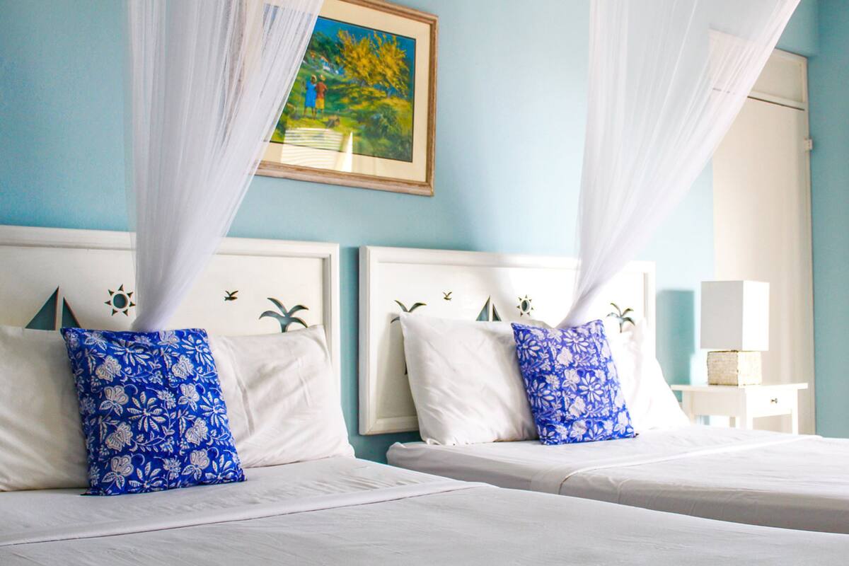 Two beds with white linens and blue floral pillows, a painting on the wall, and a white nightstand with a lamp.