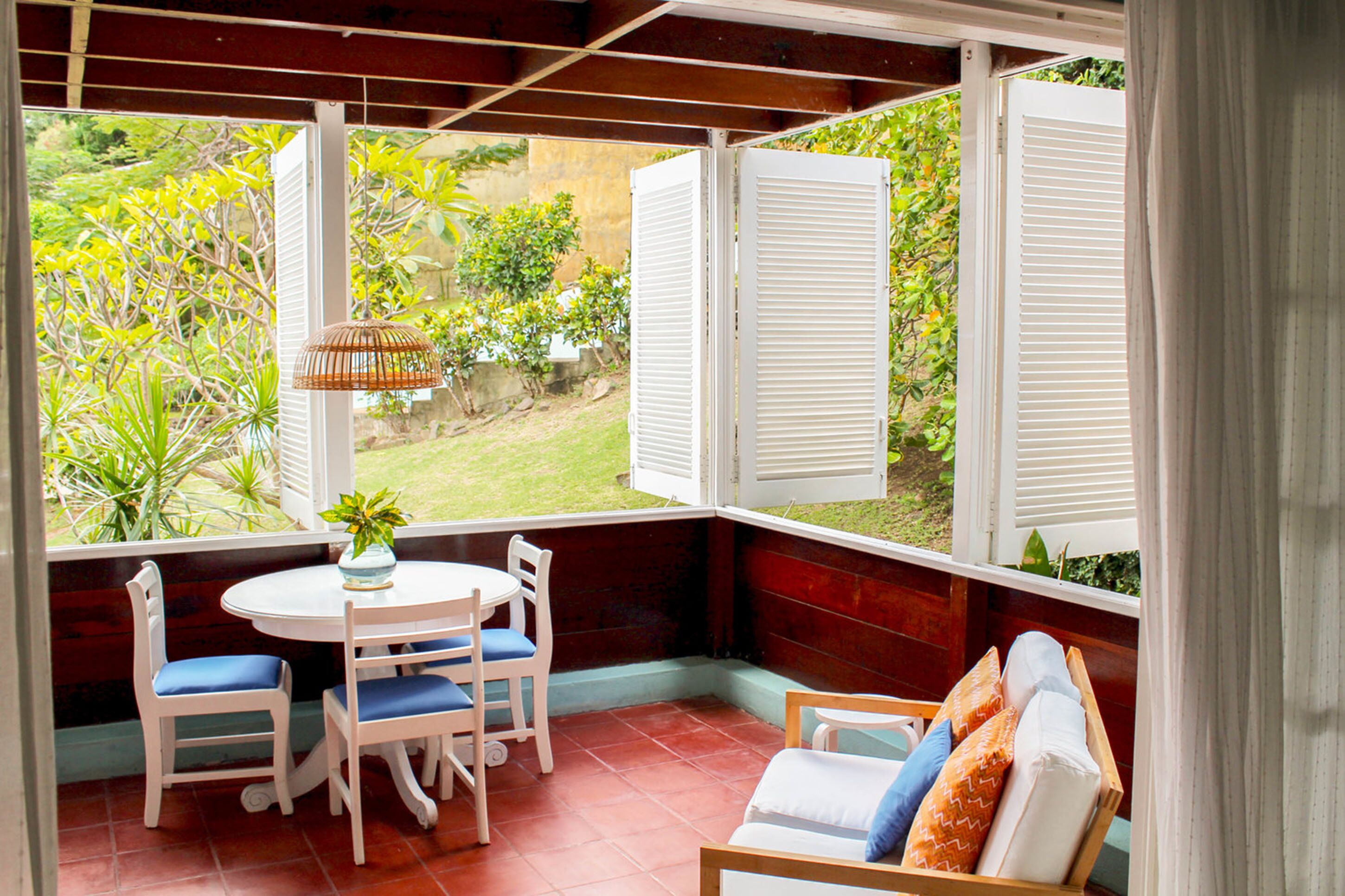 A covered patio with a table and chairs, a cushioned bench, and a view of greenery through open shutters.