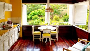 A kitchen with a window view of a garden, wooden flooring, and white furniture. - Sugarapple Inn (Bequia Island)