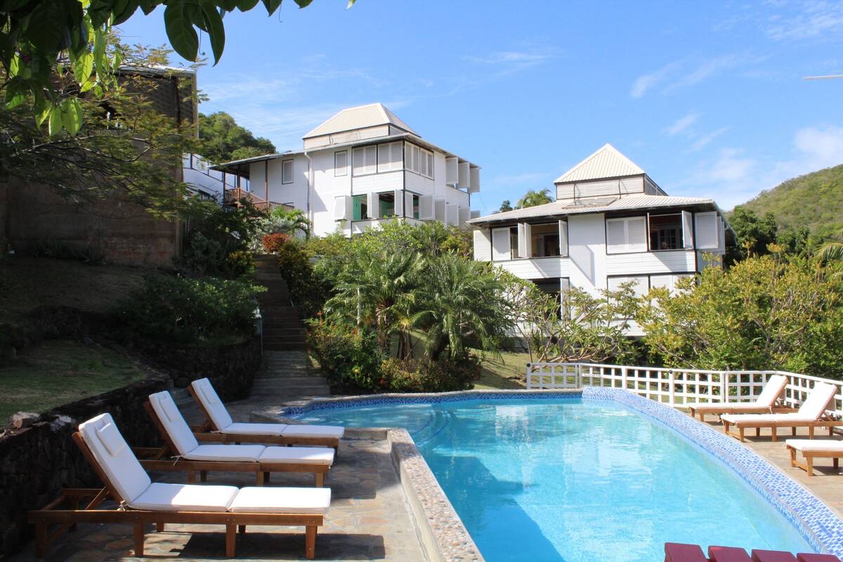 A swimming pool with lounge chairs, surrounded by a stone wall and greenery, with buildings in the background.