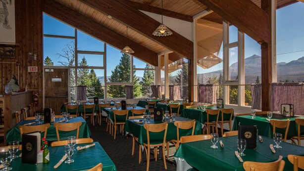 A restaurant with wooden beams, green tablecloths, and large windows offering mountain views. - Rising Sun Motor Inn and Cabins - Inside the Park (East Glacier Park)