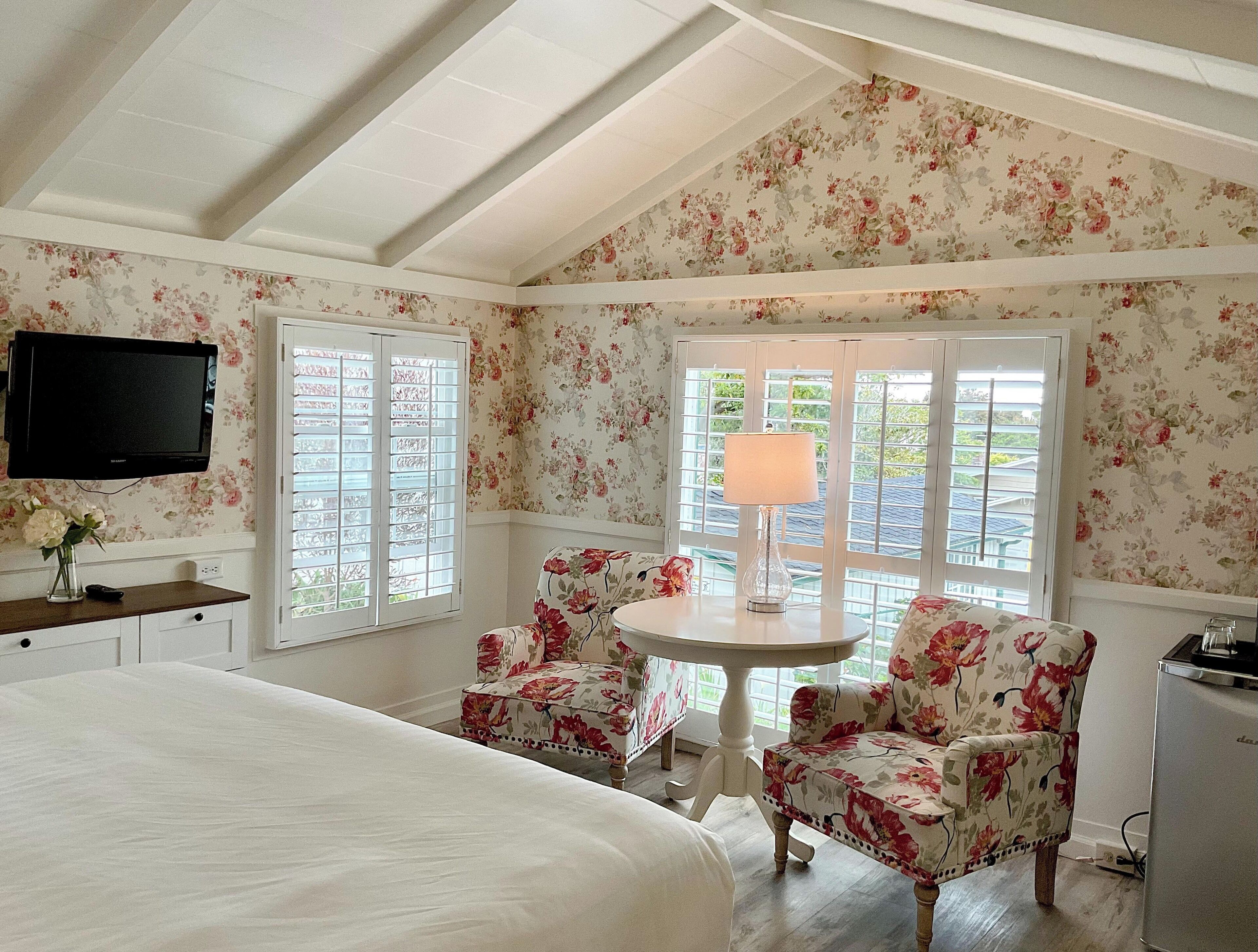 A bedroom with a bed, a TV mounted on the wall, a table with chairs, and a window with blinds.