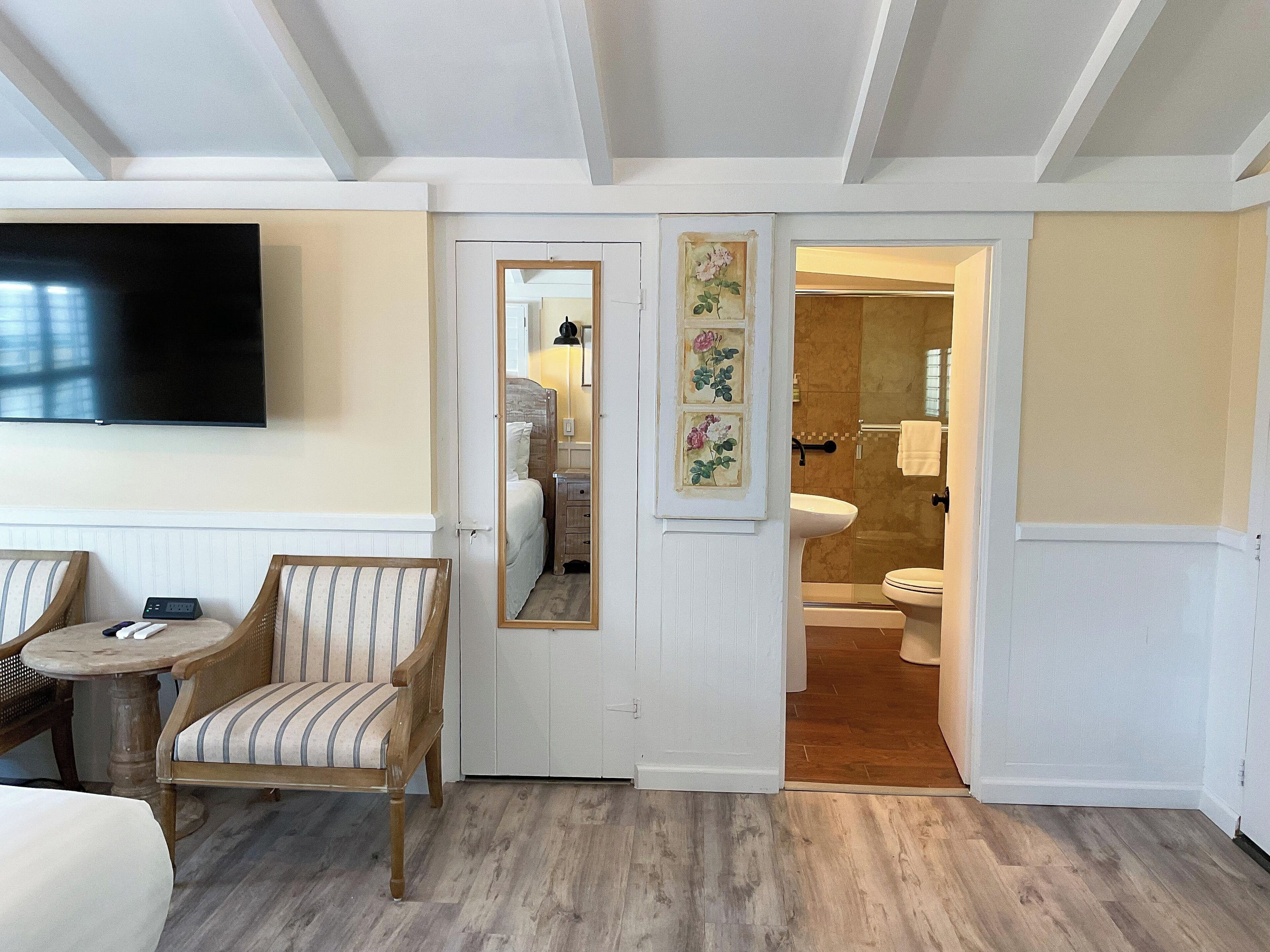 A hotel room with a TV, a bed, a chair, a small table, a mirror, and a bathroom with a toilet and sink.