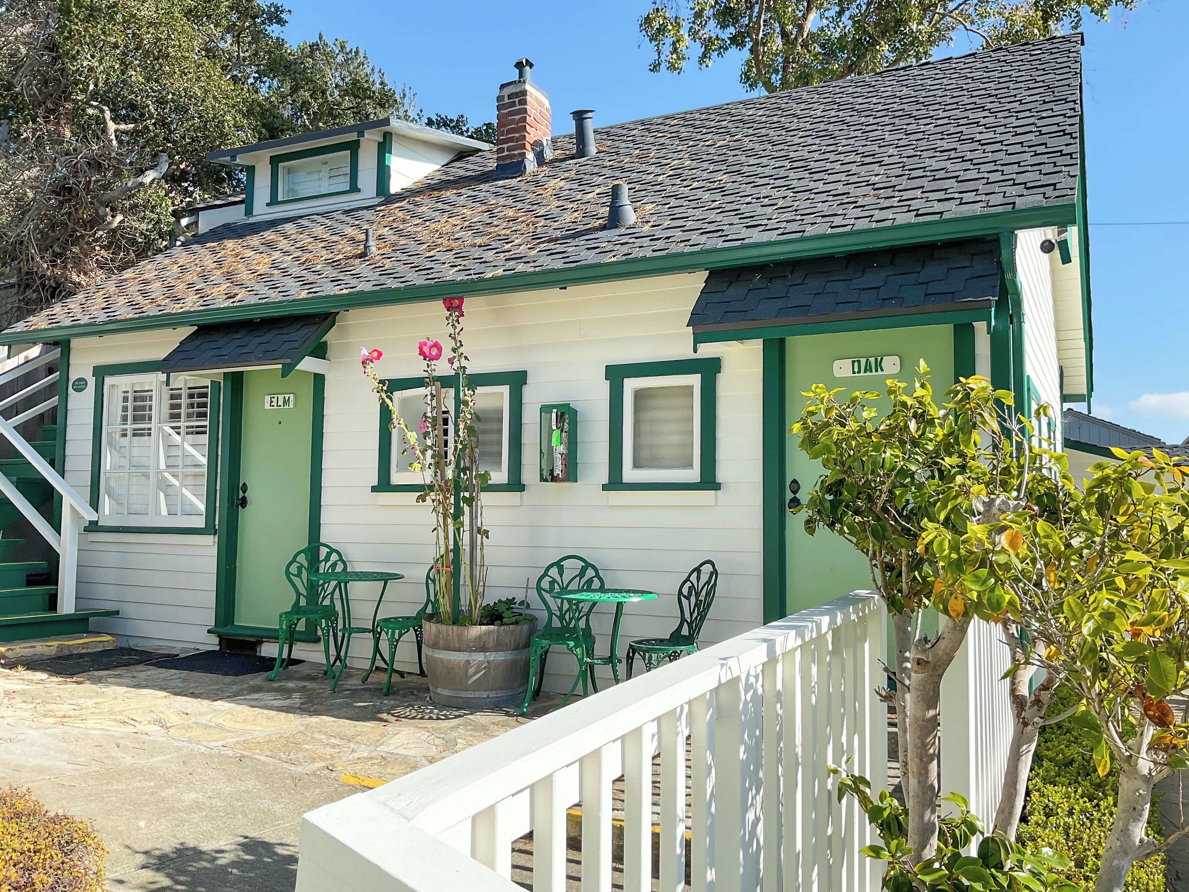 A house with a green door, a small porch with green chairs, and a white fence.