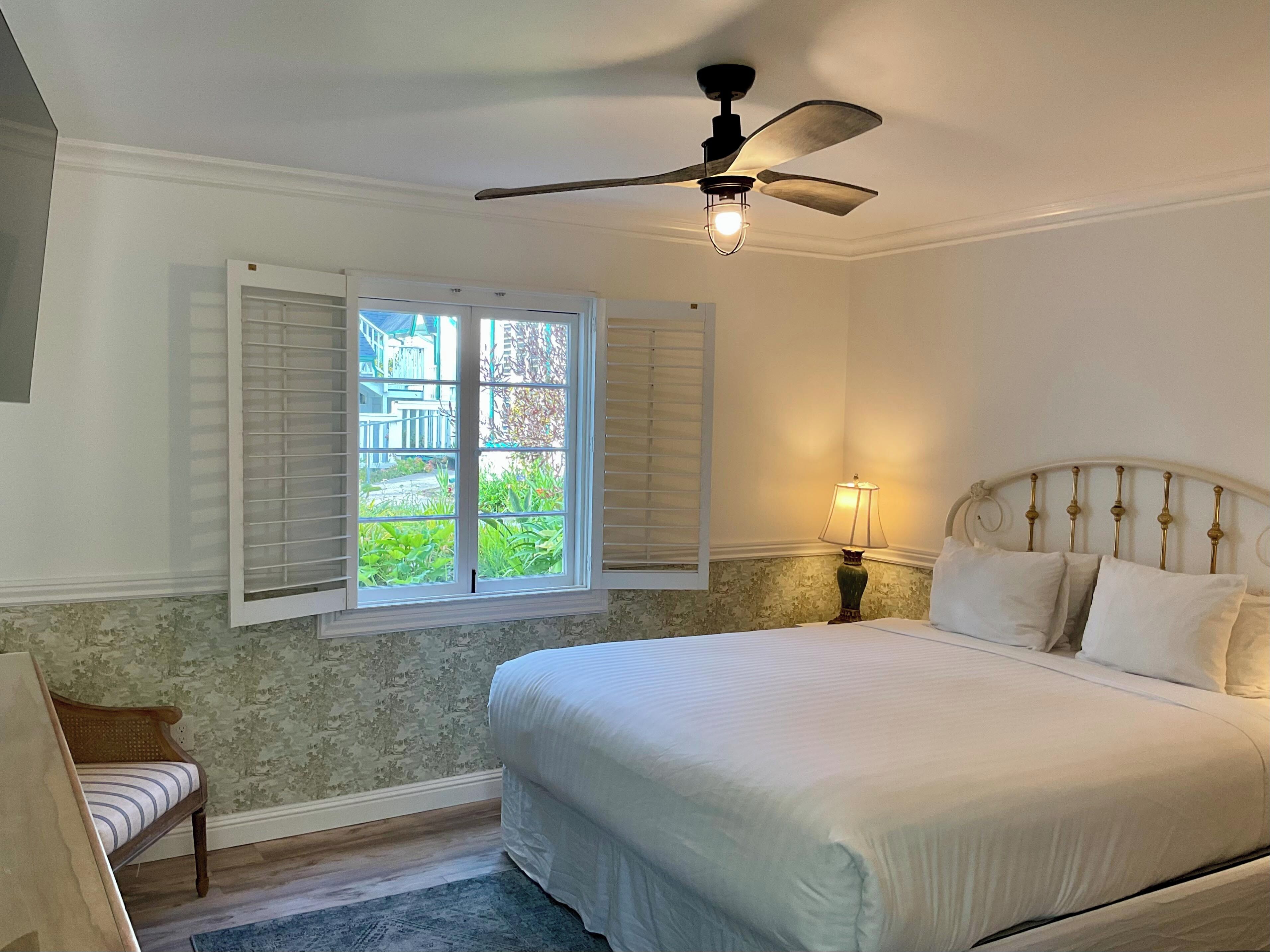 Une chambre avec un grand lit, un ventilateur de plafond, une fenêtre avec des volets, une table de chevet avec une lampe et une chaise.