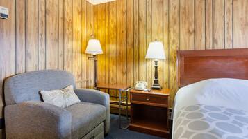 A hotel room with a wooden paneled wall, a grey armchair with a patterned pillow, a wooden desk with a telephone and lamp, and a bed with a patterned bedspread.