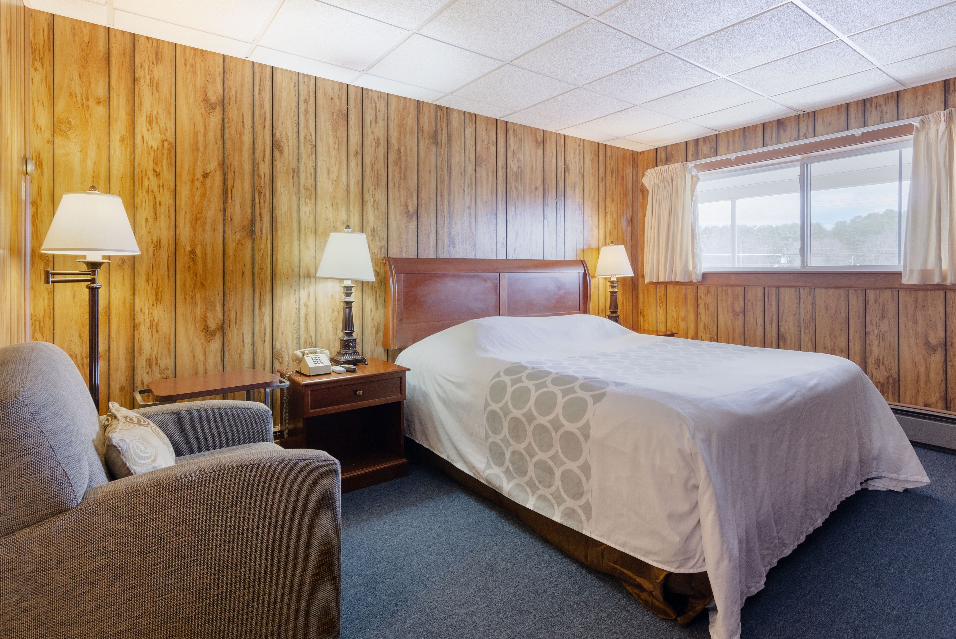 Habitación de hotel con pared de paneles de madera, una cama grande con colcha blanca, dos lámparas de noche, una mesita de noche de madera, una silla y una ventana con cortinas.