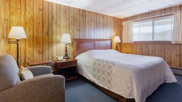 Habitación de hotel con pared de paneles de madera, una cama grande con colcha blanca, dos lámparas de noche, una mesita de noche de madera, una silla y una ventana con cortinas.
