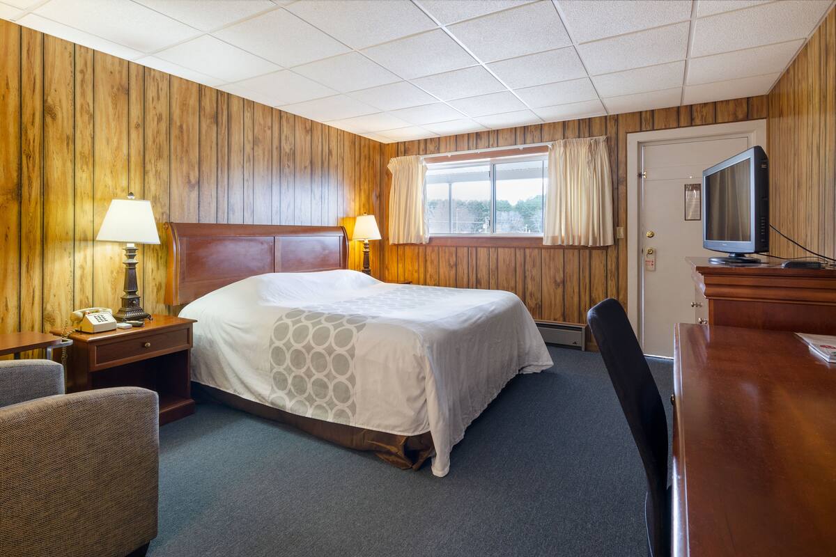 Une chambre d’hôtel avec un mur lambrissé en bois, un grand lit recouvert d’une courtepointe blanche, deux lampes de chevet, un téléviseur posé sur une table en bois et une chaise.