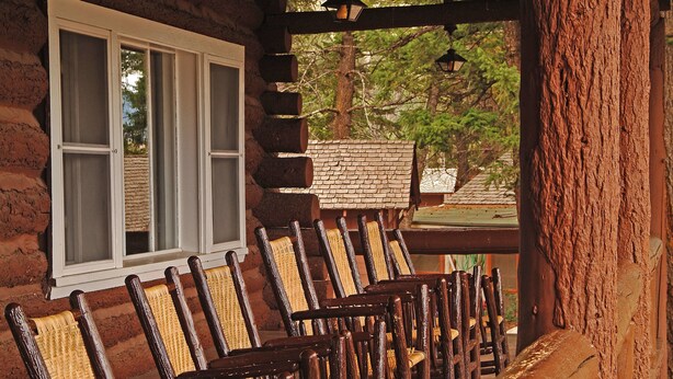 A row of wooden chairs with woven seats and backs, arranged in front of a log cabin with a window and a view of trees. - Roosevelt Lodge & Cabins - Inside the Park (Yellowstone National Park)