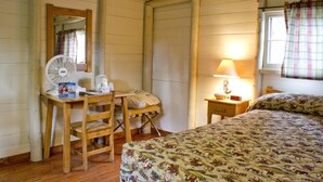 A room with a wooden desk, a chair, a bed with a floral bedspread, a standing fan, a bedside table with a lamp, and a window with curtains. - Roosevelt Lodge & Cabins - Inside the Park (Yellowstone National Park)