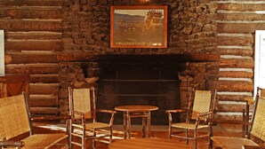A rustic interior with a stone fireplace, wooden furniture, and a framed picture of a historic scene. - Roosevelt Lodge & Cabins - Inside the Park (Yellowstone National Park)