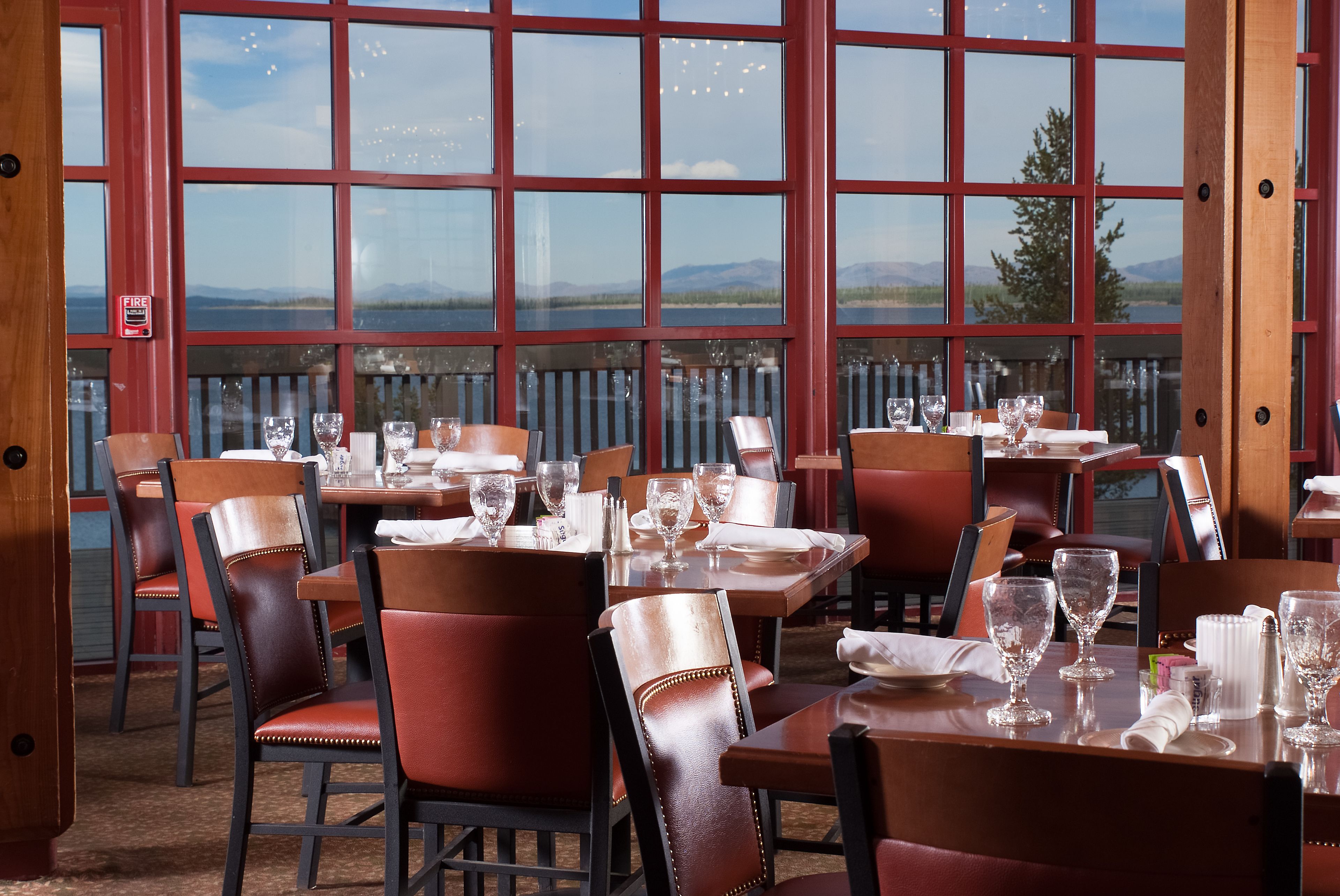 A restaurant with tables set for dining, large windows offering a view of a lake and mountains, and a fire alarm visible on the wall.