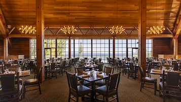 A spacious dining area with multiple tables and chairs, large windows offering a view of the outdoors, and wooden beams on the ceiling.