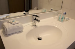 Ett hotellbadrum med en vit handfat, en spegel och hygienartiklar på hyllan.