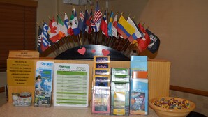 A hotel reception desk with a display of international flags, a 'Luke Guest Services' sign, and brochures for tours and events. - Jolly Roger Hotel (Marina del Rey)