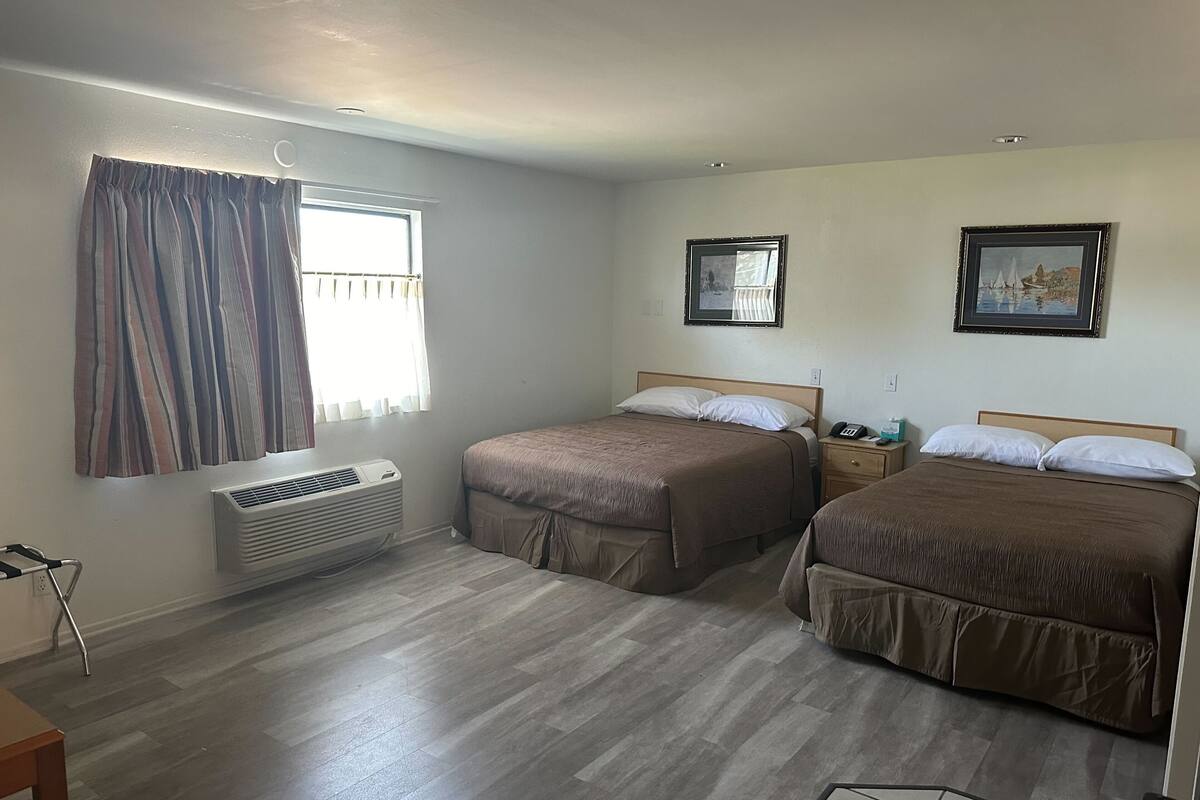 A hotel room with two beds, a window with curtains, a bedside table with a phone and a lamp, and a wall-mounted air conditioner.