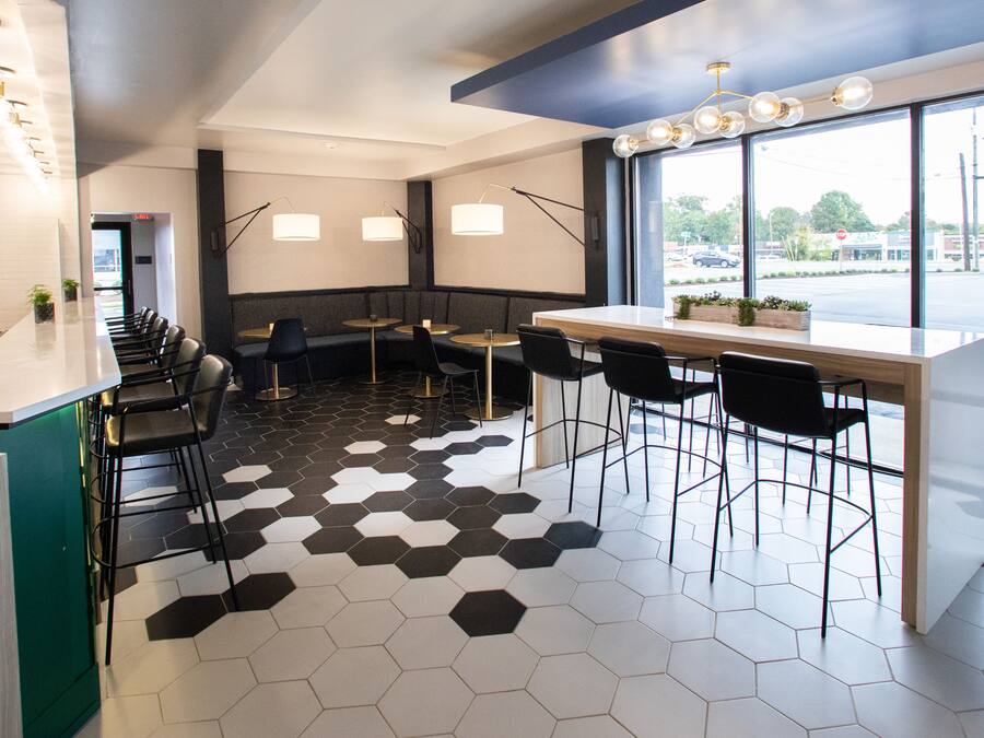Interior moderno de restaurante con una larga barra, piso con patrón hexagonal blanco y negro y grandes ventanales que ofrecen vistas a la calle.