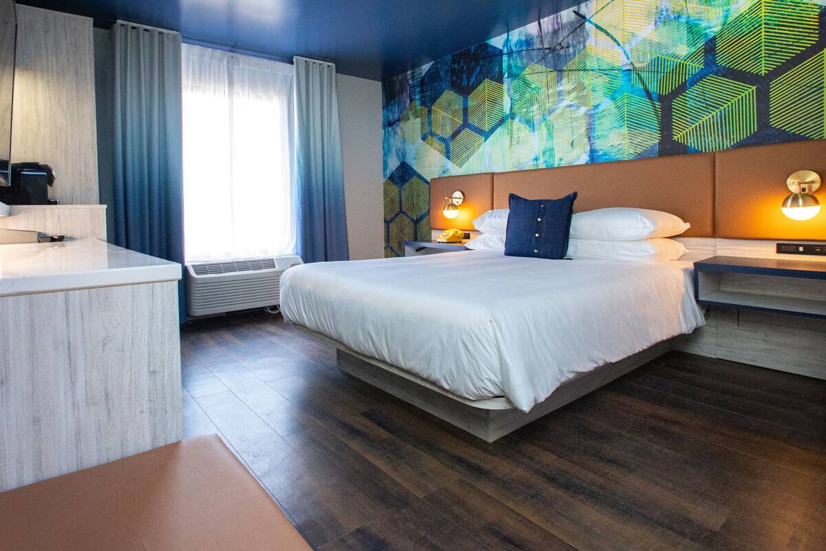 A modern hotel room with a bed, bedside tables, a desk, and a large window with curtains.
