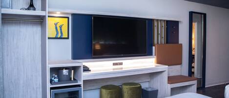 A modern hotel room with a large flat-screen TV mounted on the wall, a bed with white bedding, a small seating area with a cushioned bench, and a minibar with a bottle of water.