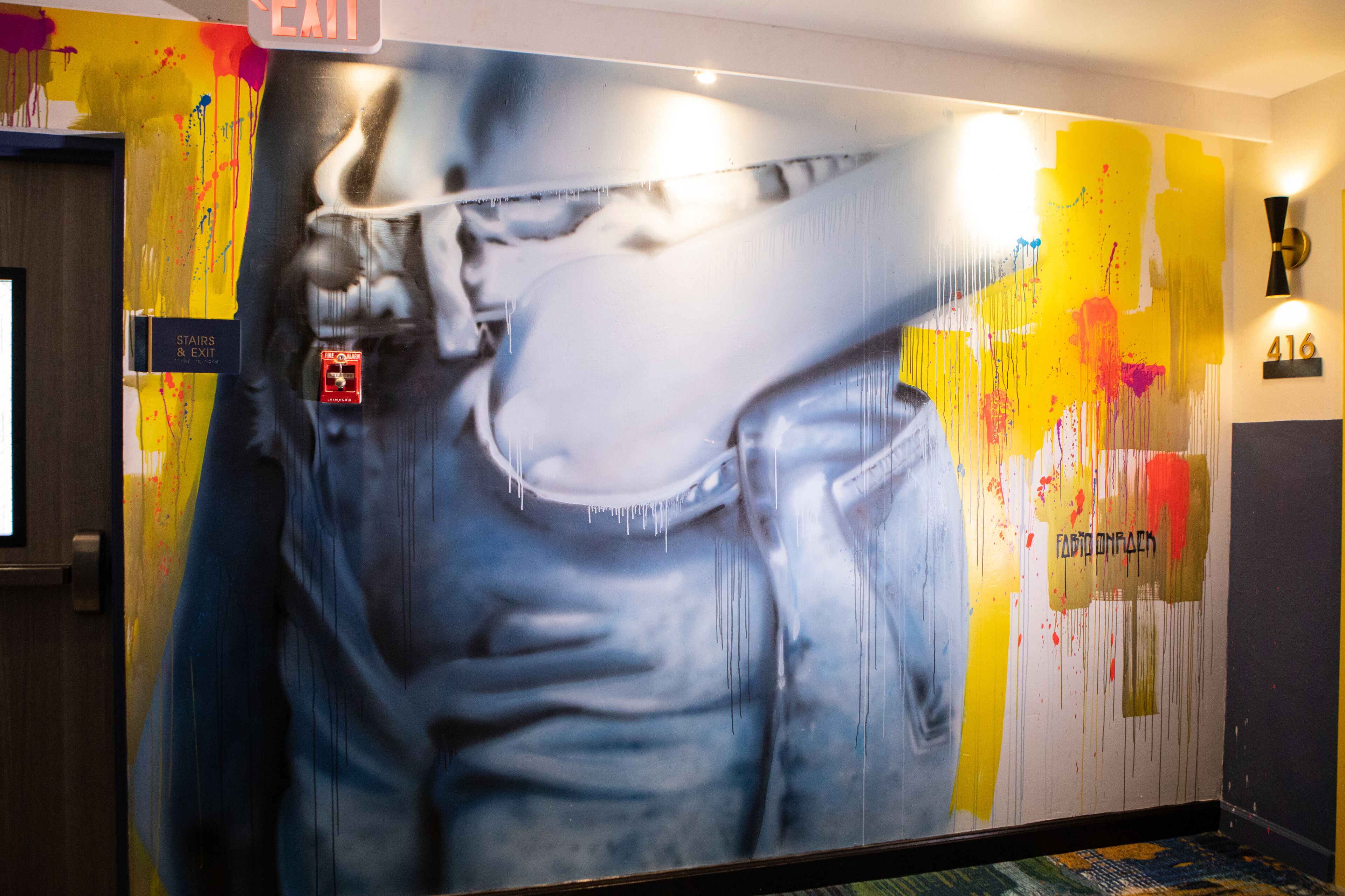 A wall with a large mural of a person wearing sunglasses and a cap. There is a sign above the mural that says "EXIT" and another sign with the number "416".