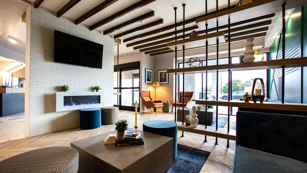 A modern living room with a fireplace, a flat-screen TV mounted on a brick wall, and a large window offering a view of the outdoors. - Hotel Denim (Greensboro)