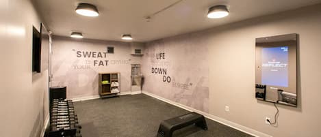 A gym with weight racks, a water dispenser, motivational wall quotes, and a digital screen displaying a fitness program.