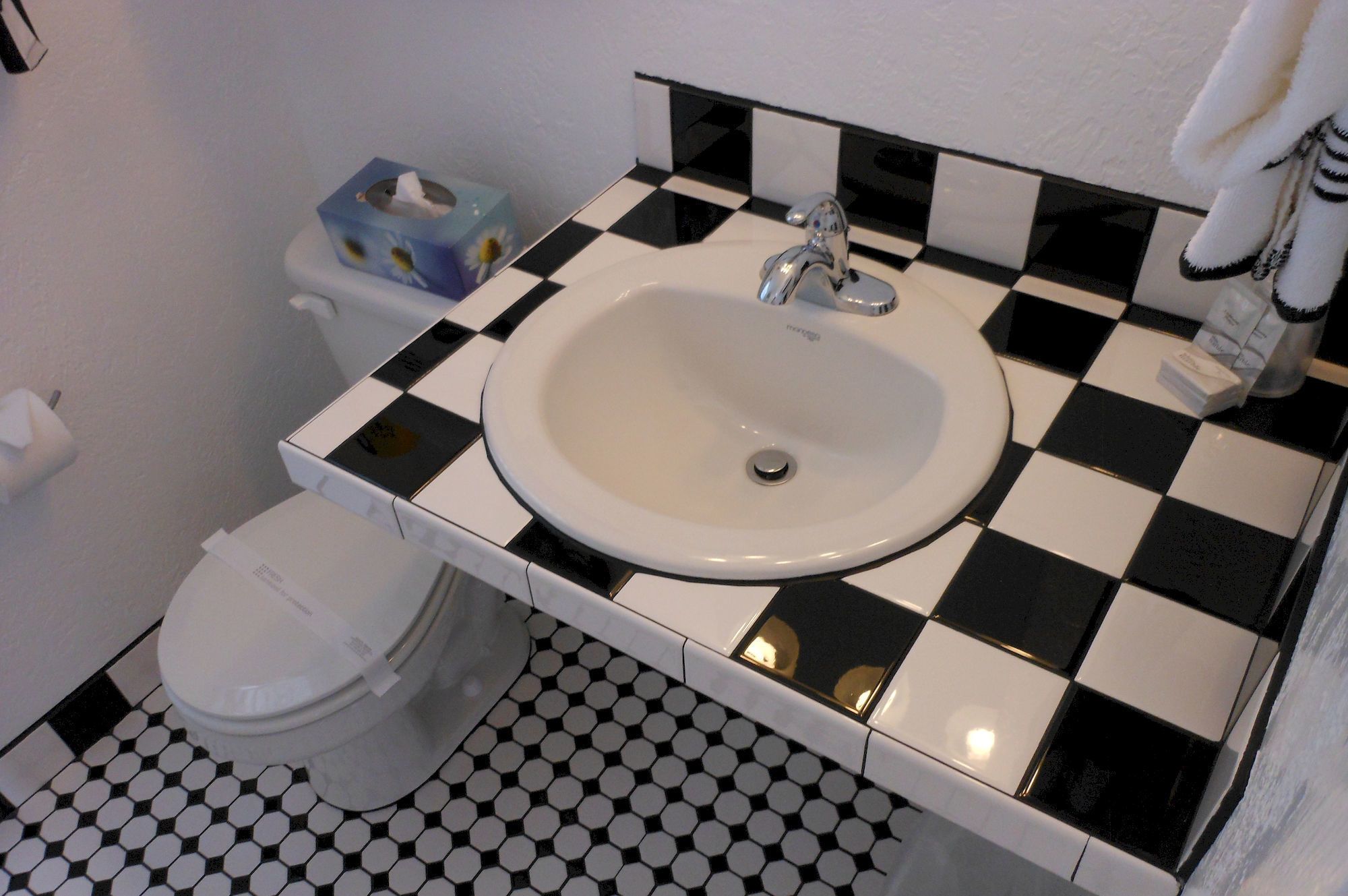 Casa de banho com uma pia branca, azulejos a preto e branco e sanita. Existe também uma caixa de lenços de papel.