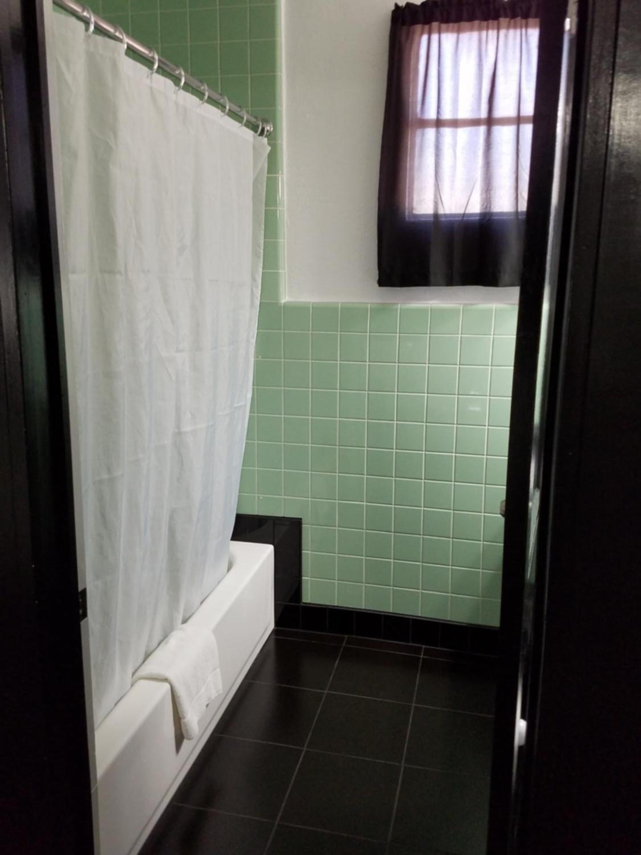 Casa de banho com banheira branca, paredes com azulejos verdes e janela com cortina preta.