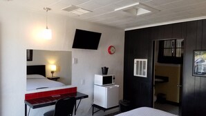 A hotel room with a bed, a desk with a chair, a microwave, a clock, and a TV mounted on the wall. - Rocket Motel (Custer)