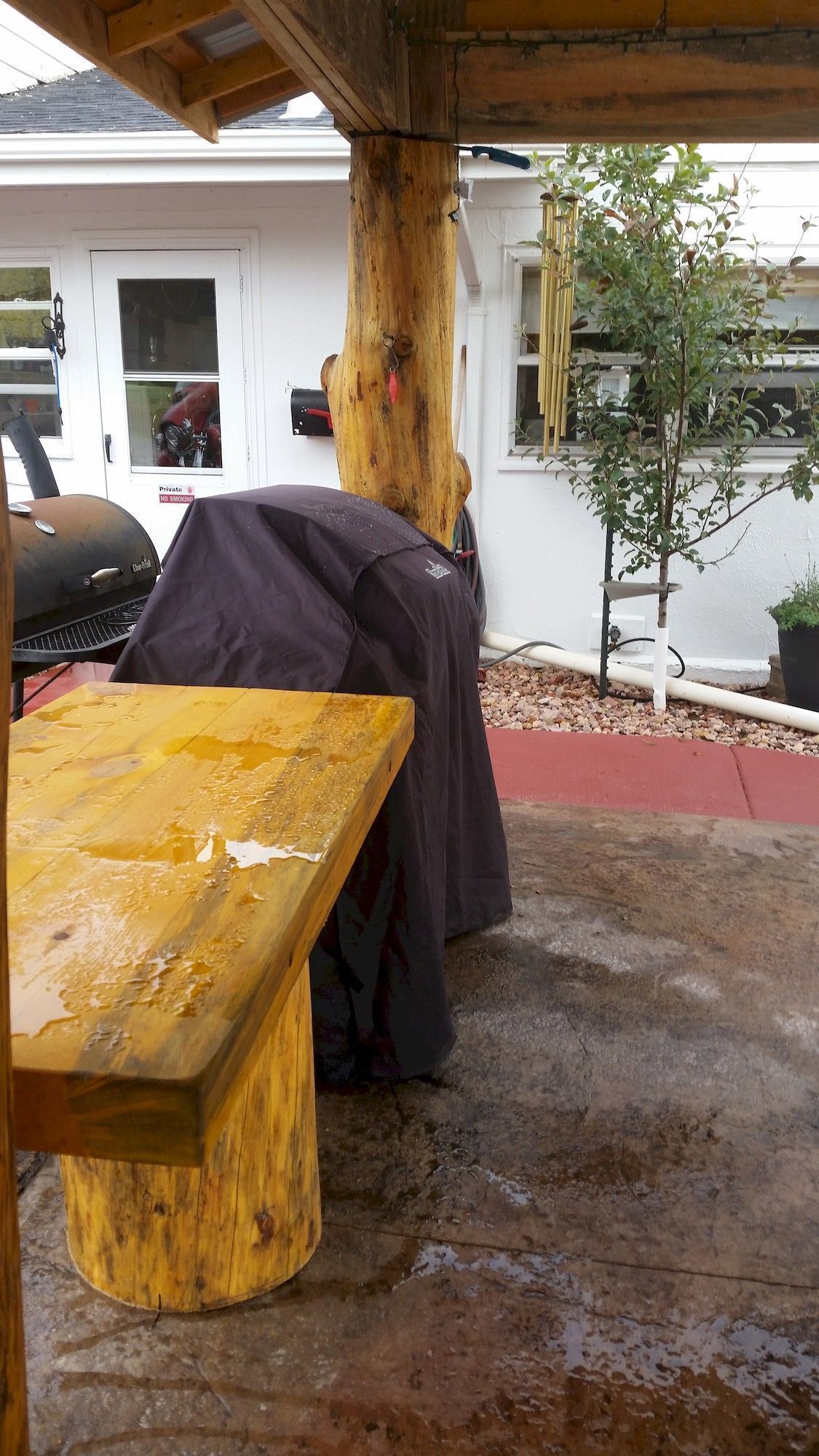 A covered outdoor seating area with a wooden bench and a covered table, a tree trunk, and a small plant.