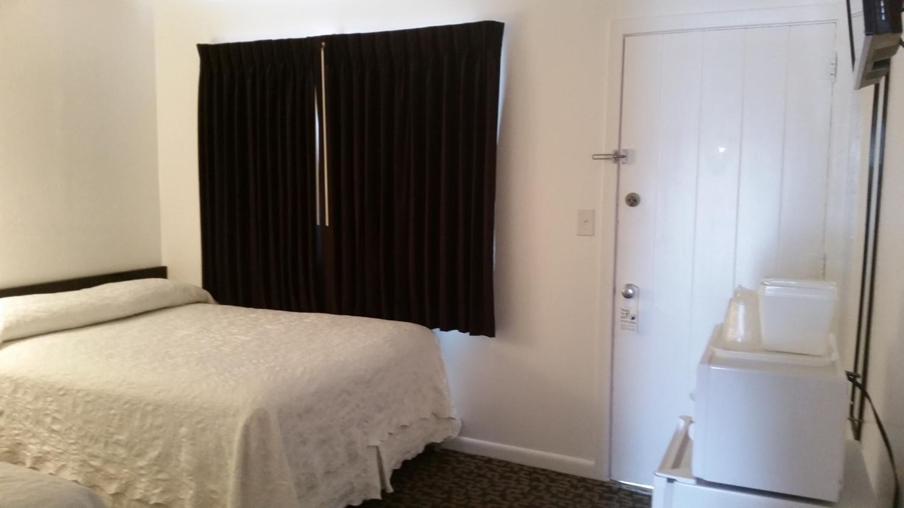 Una habitación de hotel con una cama, una puerta blanca y un refrigerador blanco.