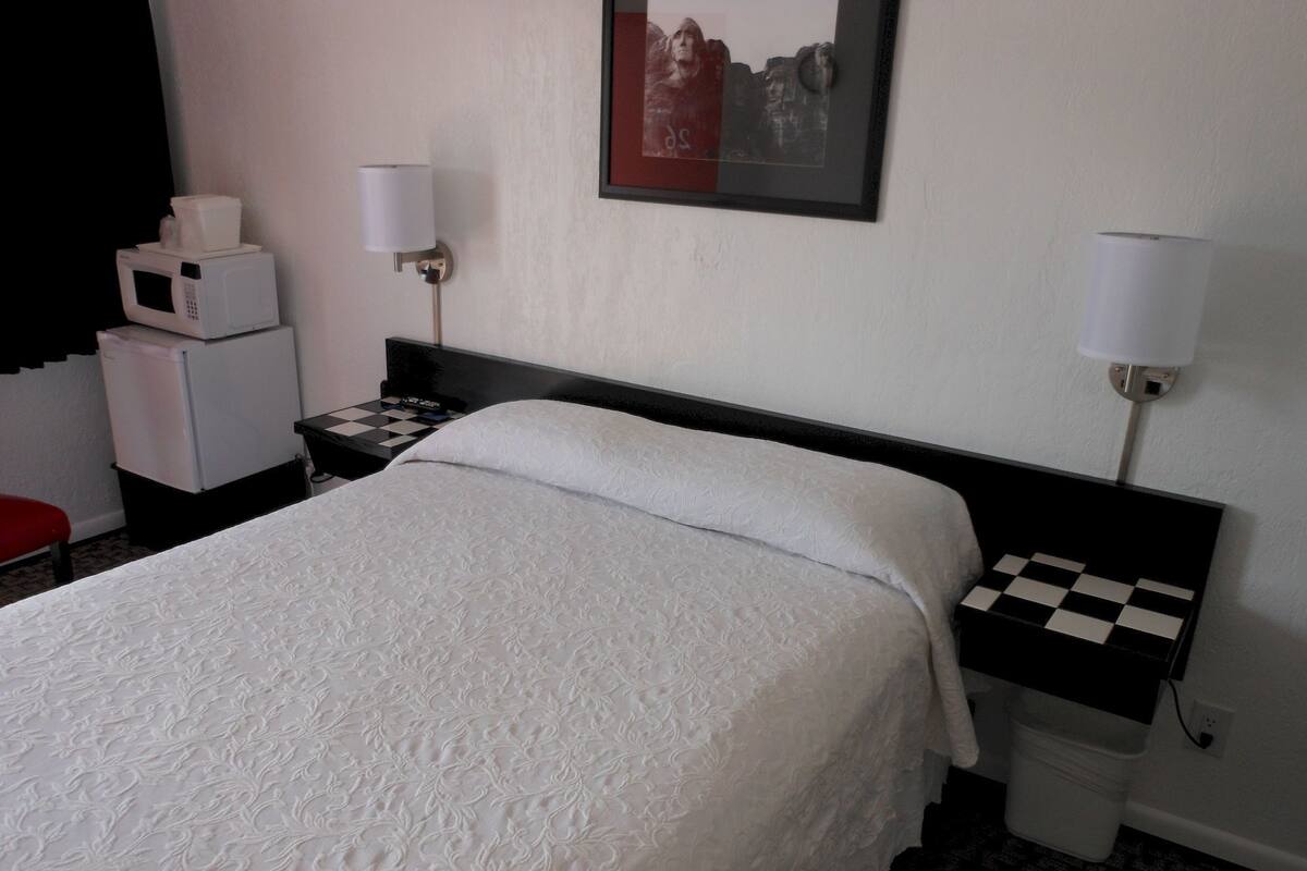 A hotel room with a bed, a nightstand, a microwave, a refrigerator, a checkered nightstand, and two wall-mounted lamps.