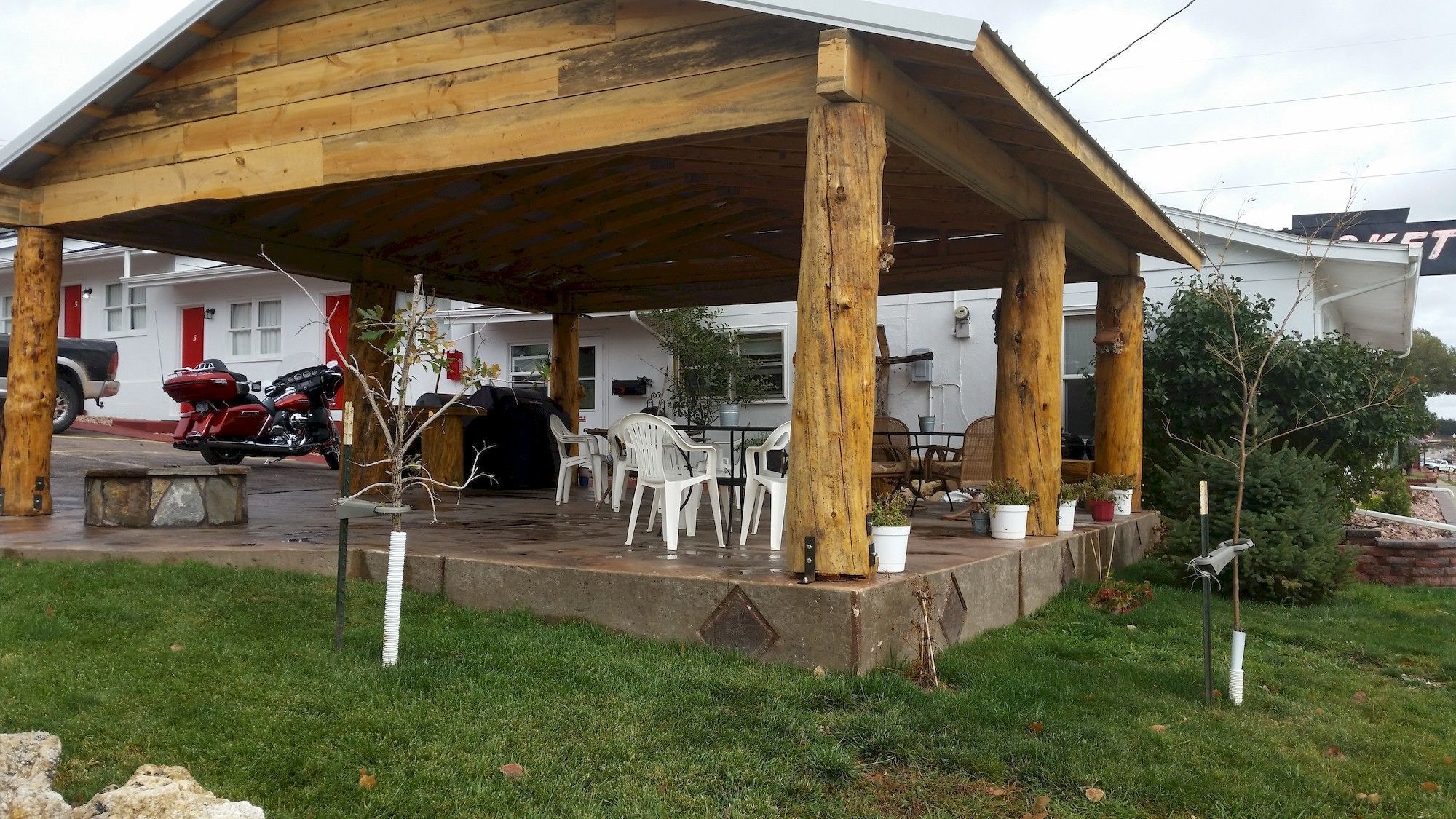 A covered outdoor seating area with concrete flooring, surrounded by potted plants and white plastic chairs.