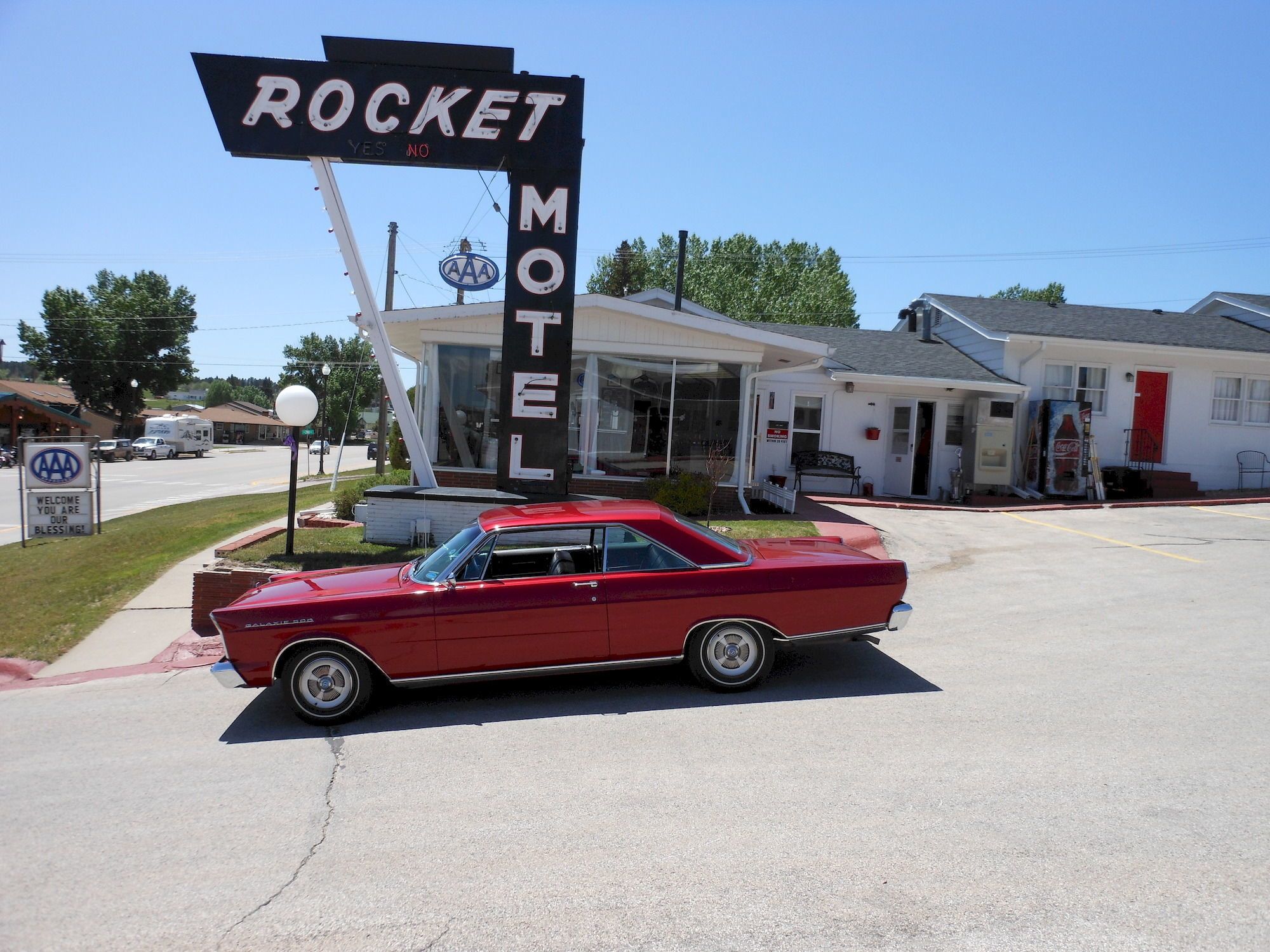 A vintage red car parked in front of a motel with a sign reading "ROCKET MOTEL."