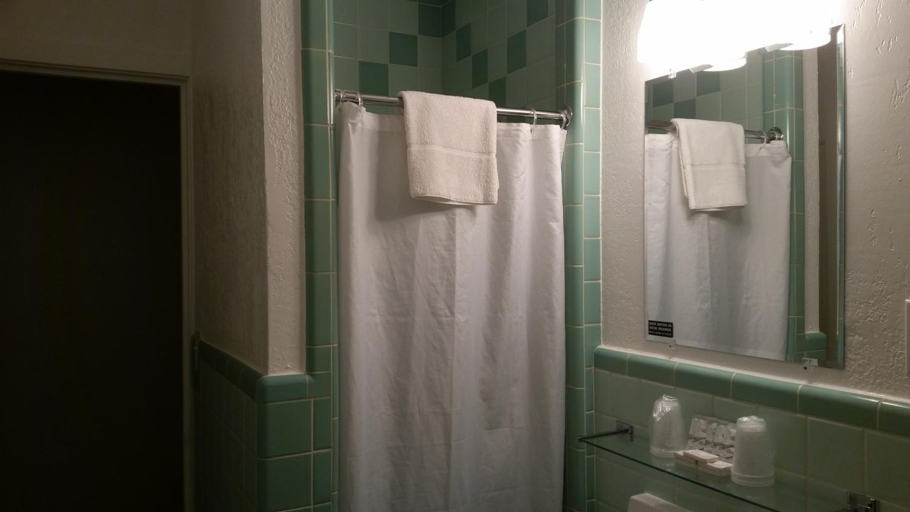 A bathroom with a white shower curtain, a glass shelf with toiletries, and a mirror reflecting the shower area.