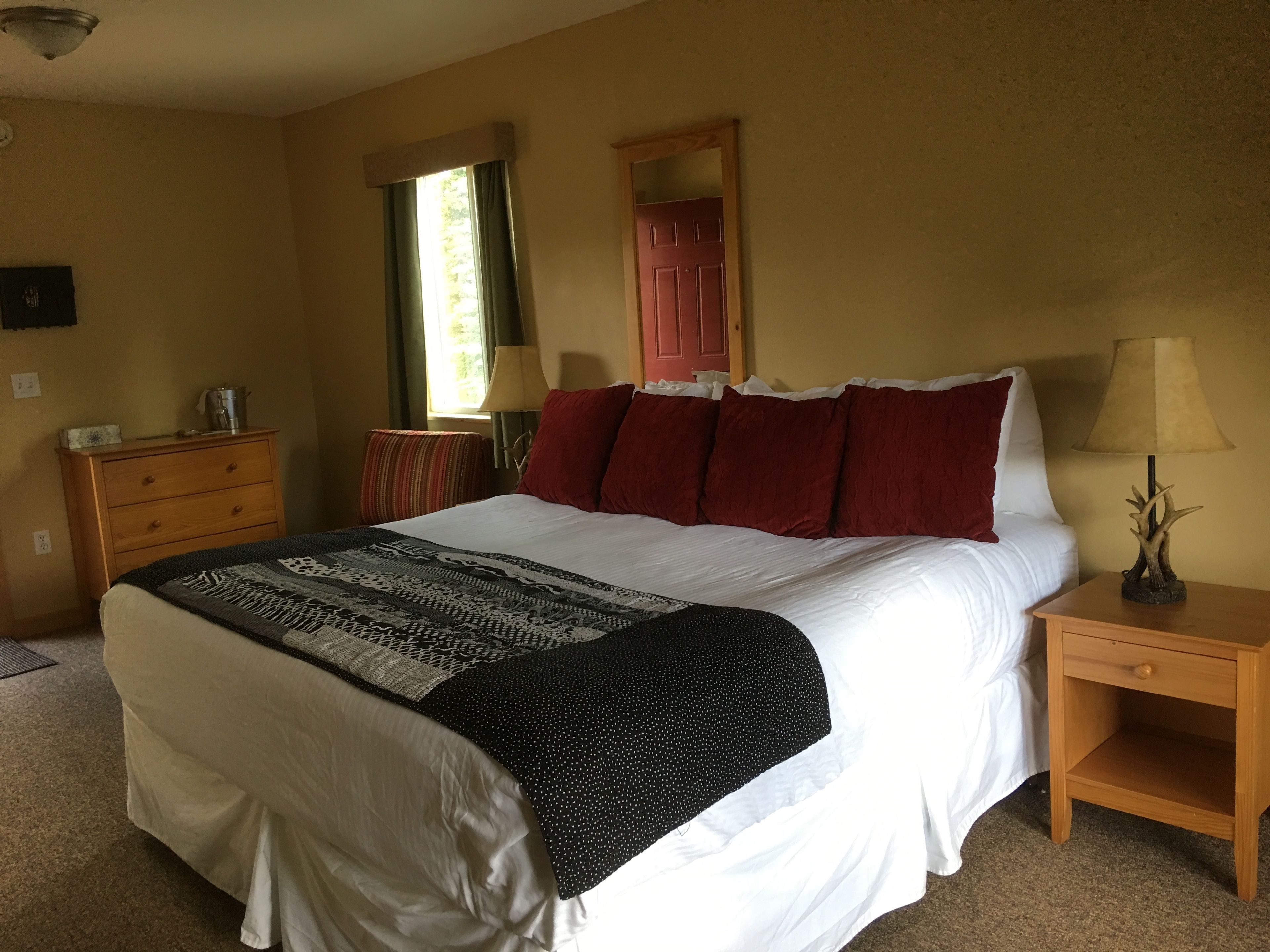 A neatly made bed with a patterned blanket, a wooden nightstand with a lamp, a dresser, and a window with curtains.