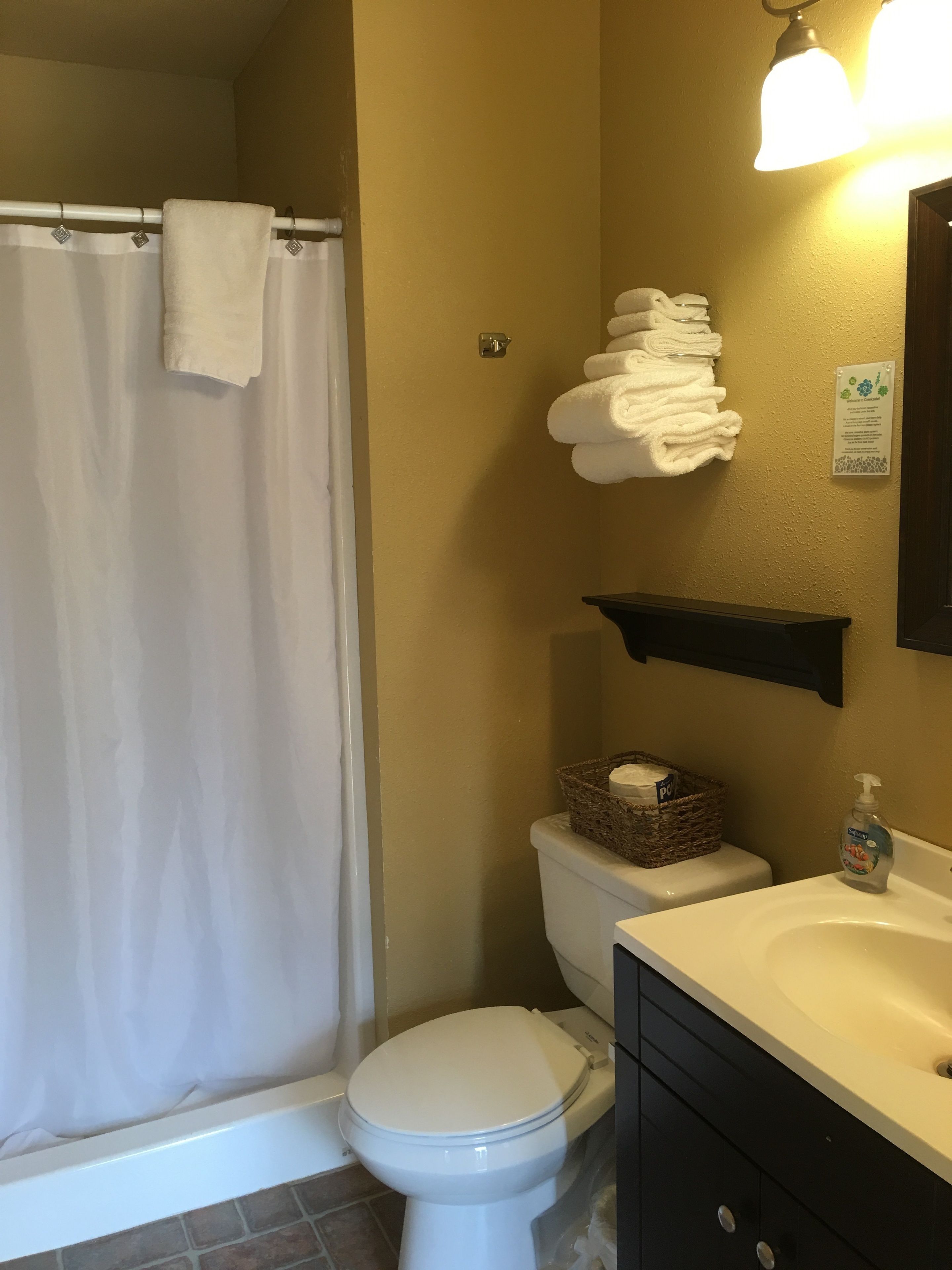 A bathroom with a shower curtain, a toilet, a sink with a soap dispenser, and a basket of toilet paper.