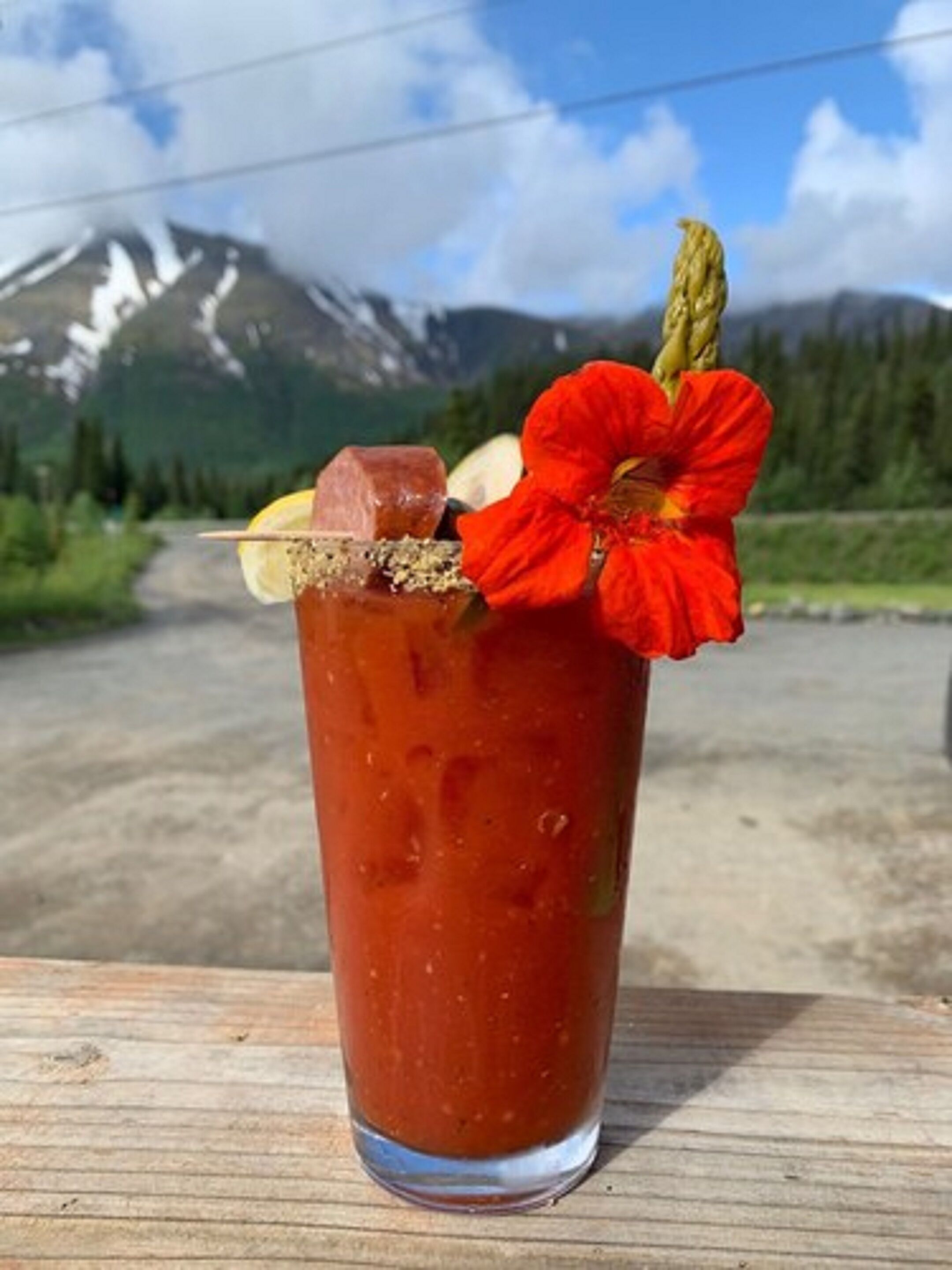 Un grand verre de Bloody Mary, décoré d’une fleur rouge, posé sur une surface en bois, avec un paysage montagneux en arrière-plan.