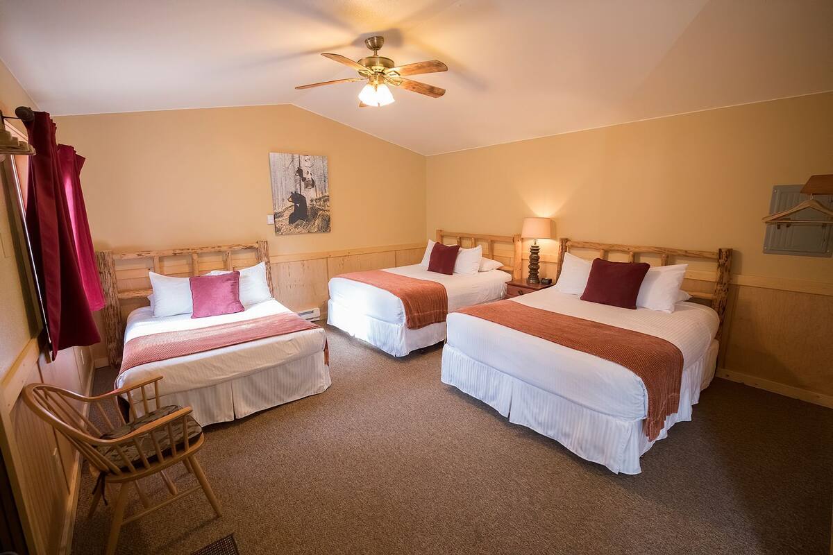 A hotel room with three single beds, a wooden chair, a ceiling fan, and a painting on the wall.