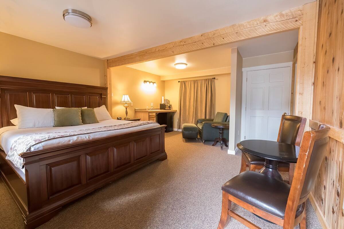 A hotel room with a large bed, a desk with a lamp, a chair, and a small table.