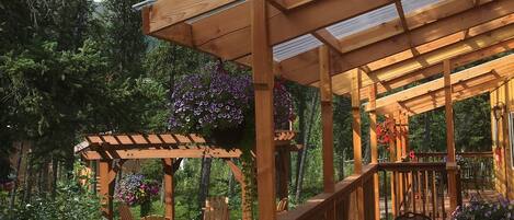 A wooden deck with a pergola and seating area, surrounded by trees and flowers.