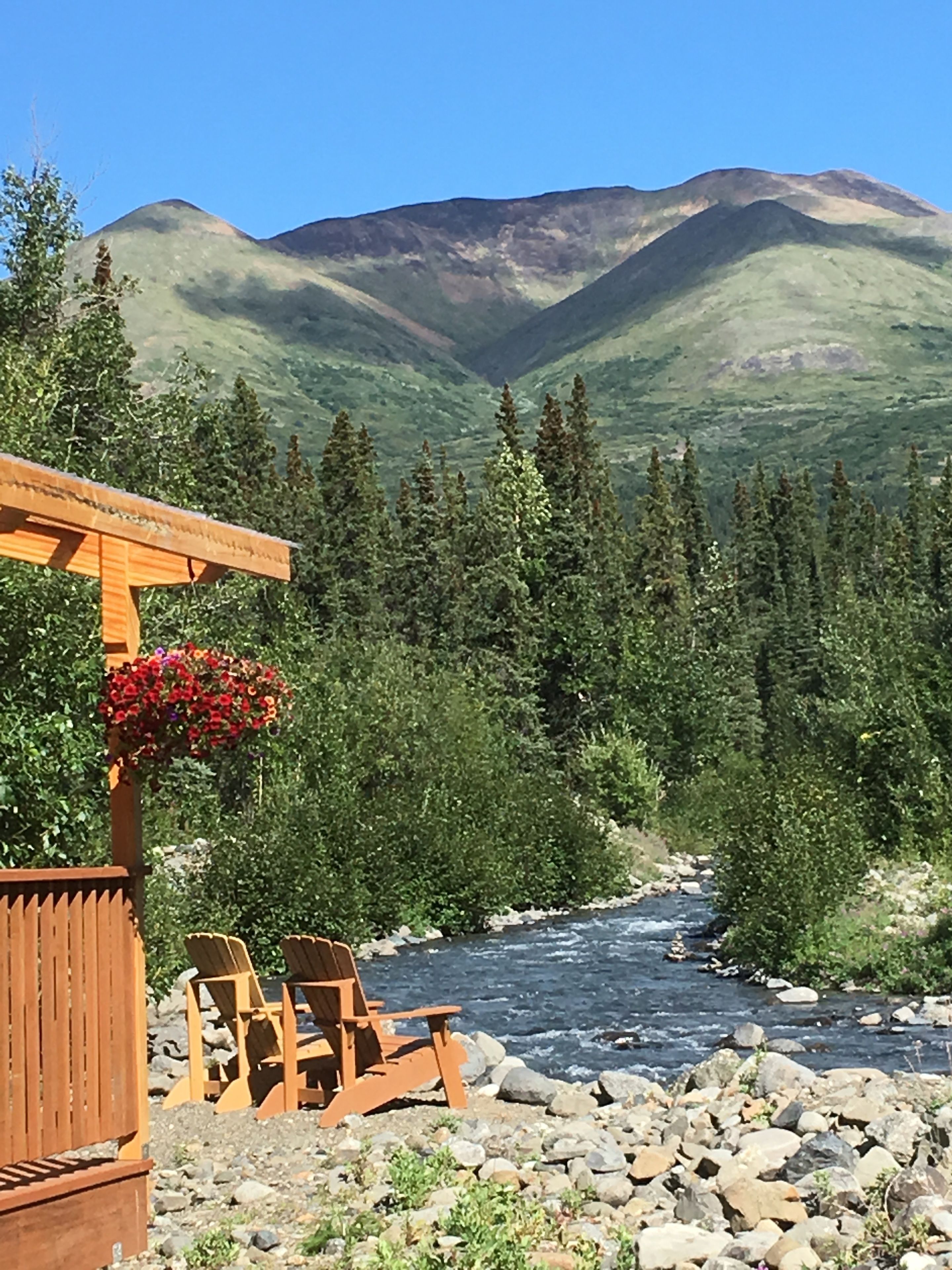 A wooden deck with chairs and a hanging flower basket overlooking a river with mountains in the background.