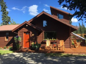 A wooden cabin with a front porch, a dining area with chairs, and a large window overlooking a forested area. - McKinley Creekside Cabins (Denali National Park)